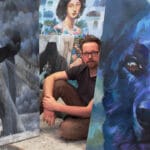 Taiteilija istuu maalauksiensa keskellä galleriassa.